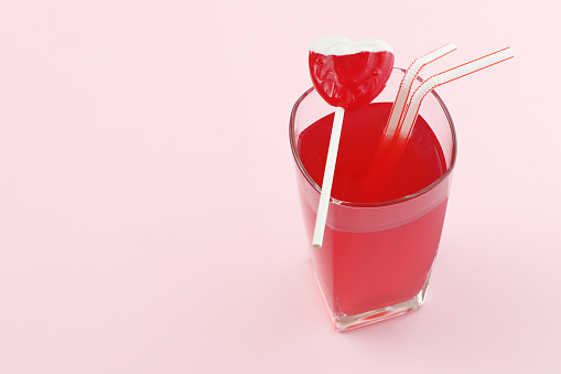 A picture of the heart shaped lollipop on top of the glass with red drink. Two straws are symbol of two people being together.